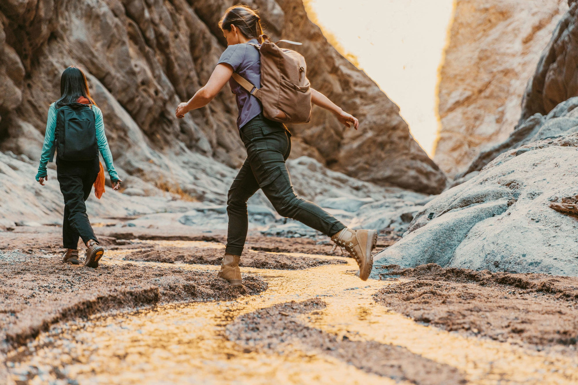Two people with backpacks hike through a narrow canyon with rocky walls and a small stream, under warm lighting.